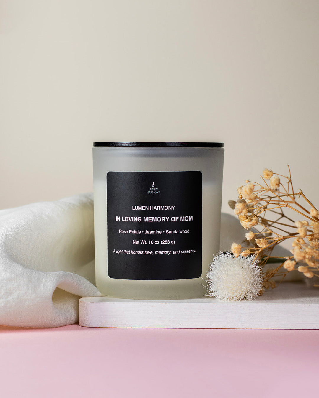 A candle called "in loving memory of Mom" over a wooden white table with tiny flowers as decoration in a neutral background
